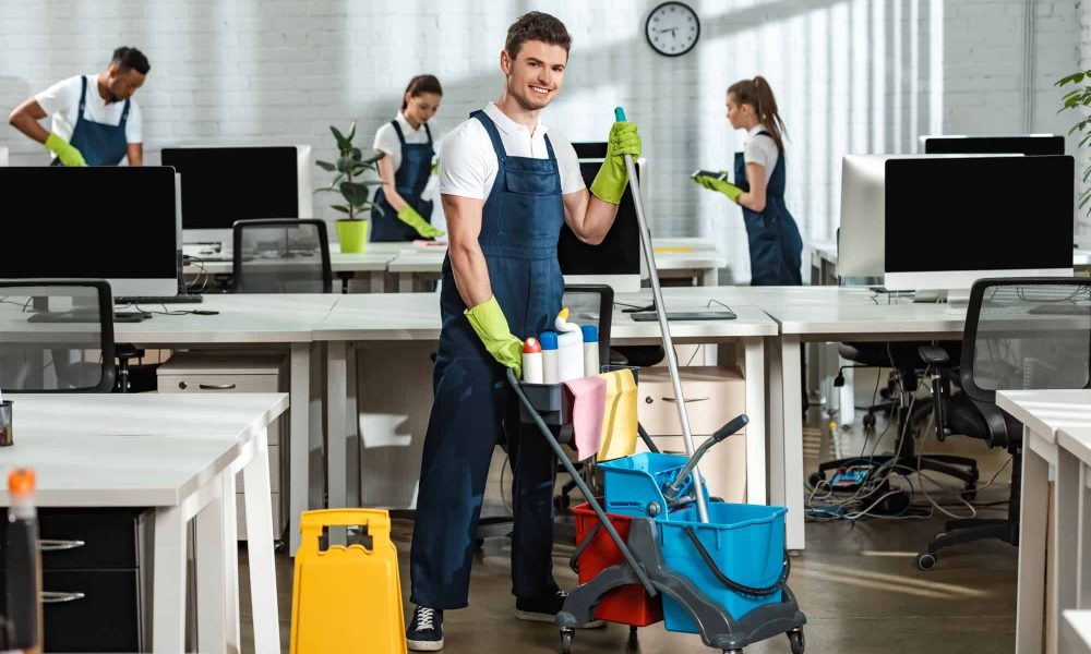 cheerful-cleaner-moving-cart-with-cleaning-supplies-near-multicultural-colleagues.jpg